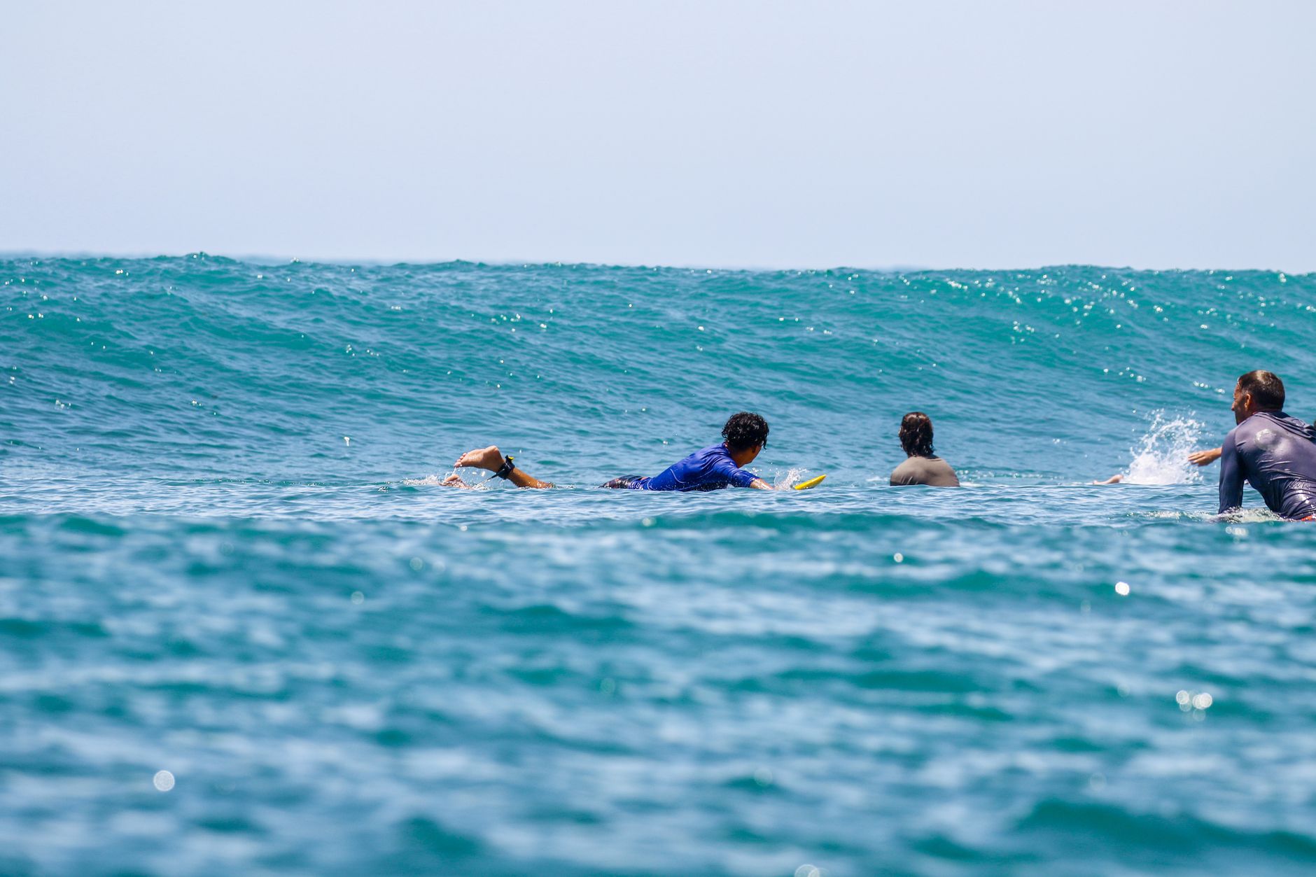Four surfers paddling on surfboards, preparing for an ocean wave on a sunny day.