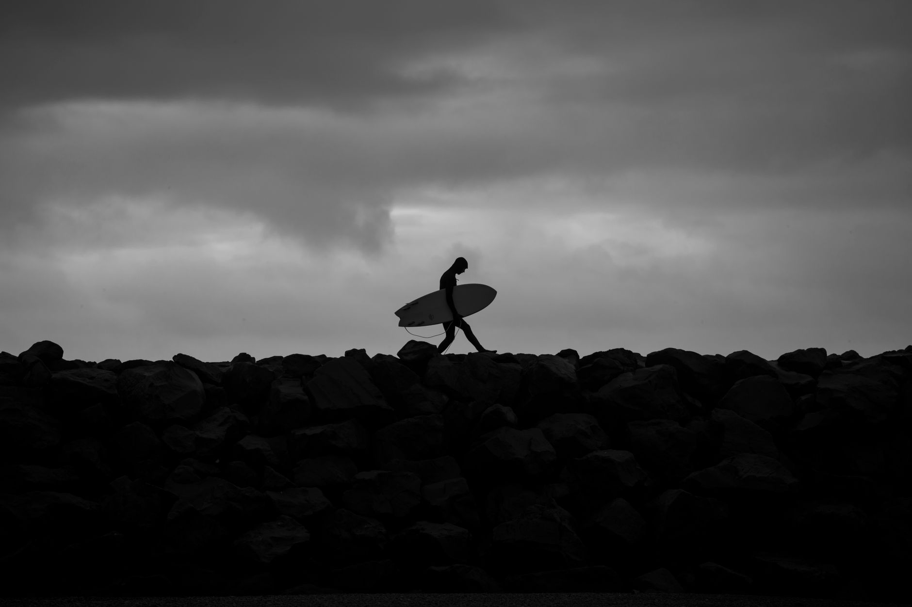 Black and white silhouette of a surfer walking along a rocky beach at dusk.