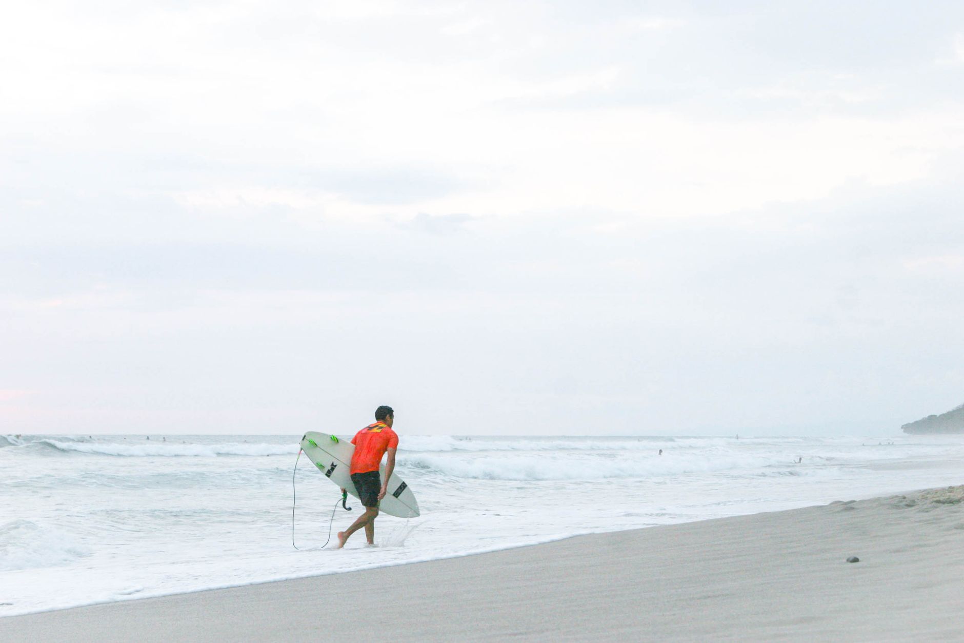 A surfer in a red shirt walks along the serene beach in Costa Rica, surfboard in hand.