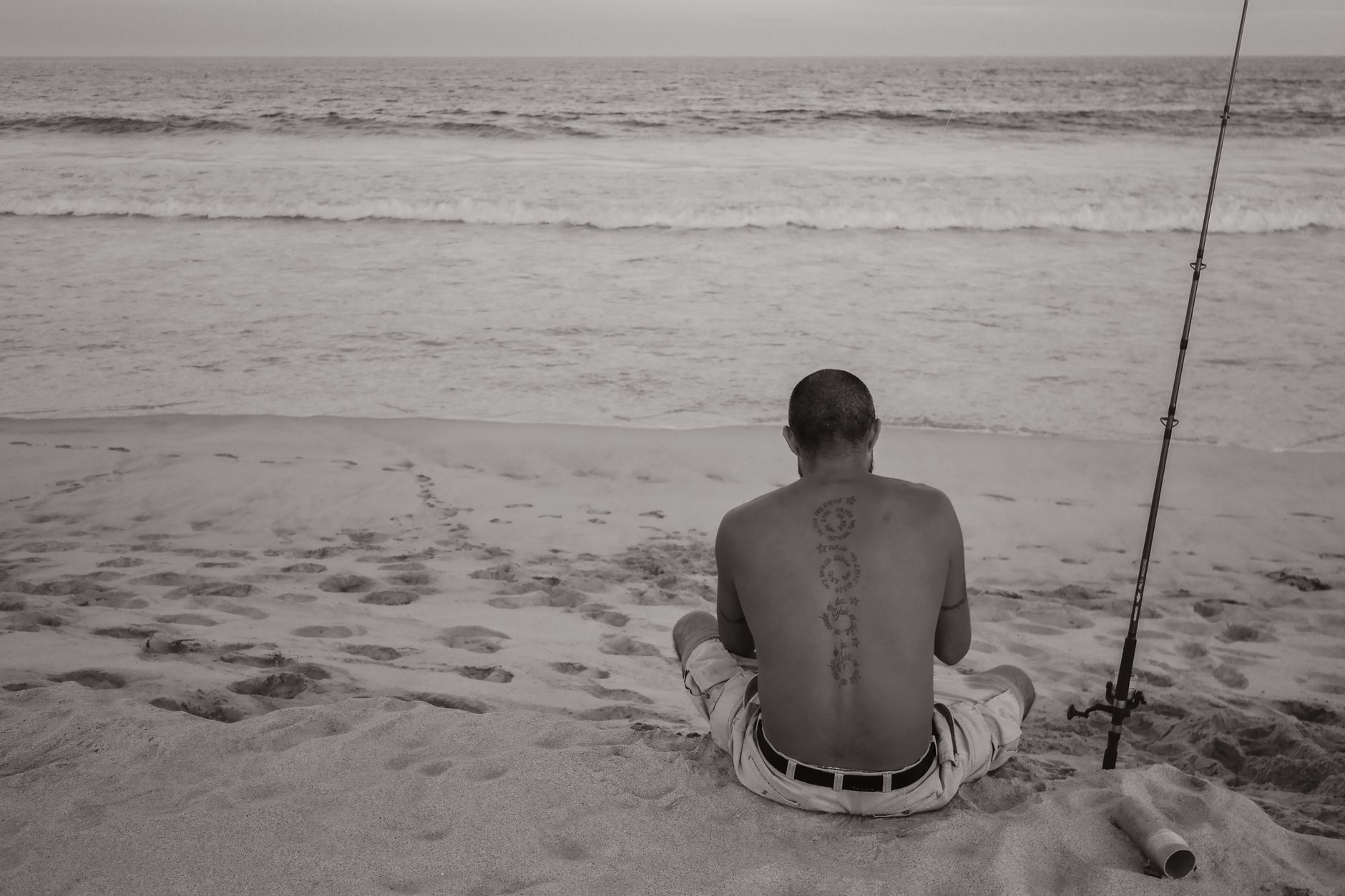 A shirtless man with tattoos fishes on a tranquil beach in San José del Cabo, Mexico, enjoying the serene ocean view.