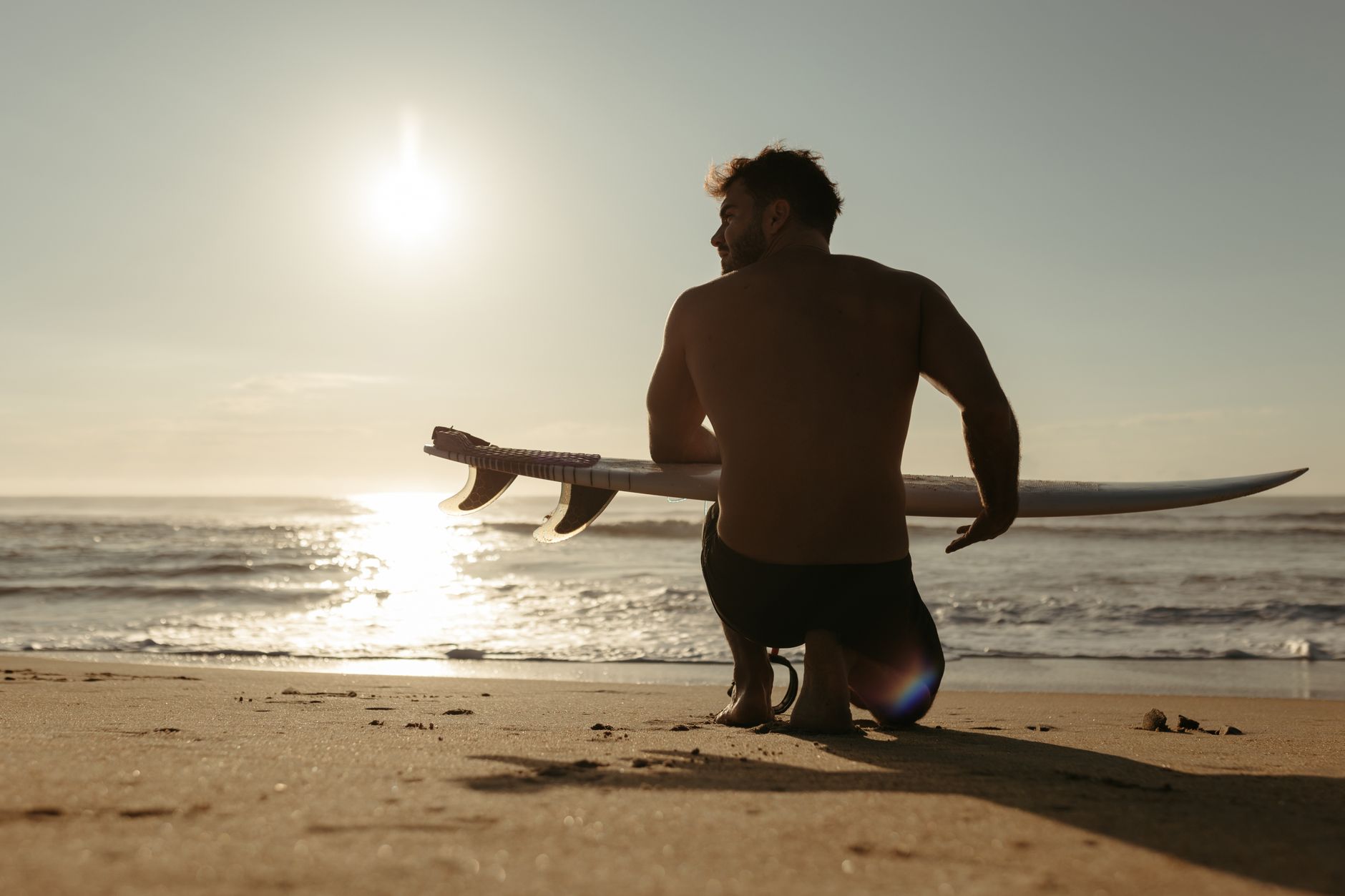 A surfer sits on a sandy beach holding a surfboard, with a stunning sunset in the background.